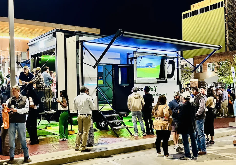 Guests gathered around a Dryvebox trailer taking swings at a night-time event lit up under the lights